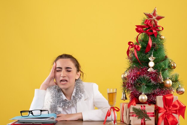 Ärztin der Vorderansicht, die hinter ihrem Tisch sitzt, der auf gelbem Hintergrund mit Weihnachtsbaum und Geschenkboxen traurig fühlt