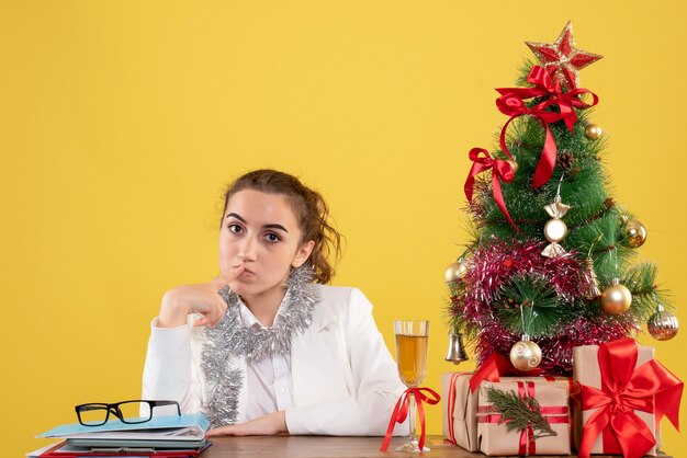 Ärztin der Vorderansicht, die hinter ihrem Tisch sitzt und auf gelbem Hintergrund mit Weihnachtsbaum und Geschenkboxen denkt