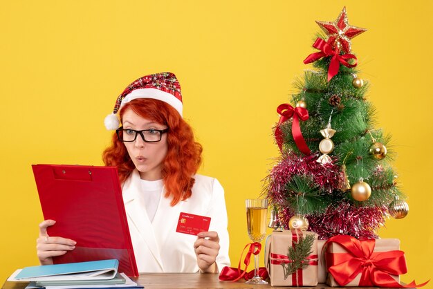Ärztin der Vorderansicht, die hinter ihrem Tisch sitzt und Bankkarte auf gelbem Hintergrund mit Weihnachtsbaum und Geschenkboxen hält