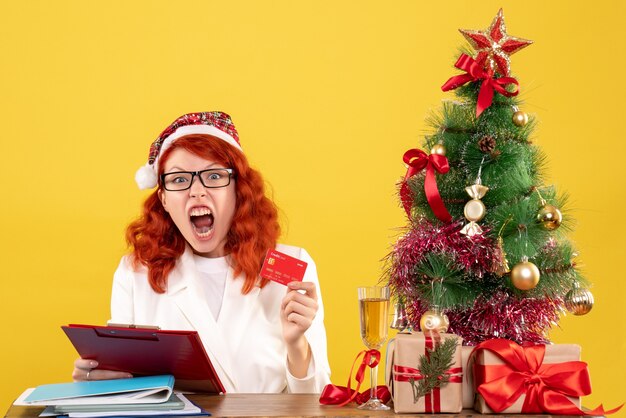 Ärztin der Vorderansicht, die hinter ihrem Tisch sitzt und Bankkarte auf gelbem Schreibtisch mit Weihnachtsbaum und Geschenkboxen hält