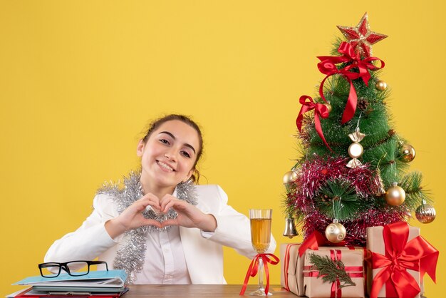 Ärztin der Vorderansicht, die hinter ihrem Tisch sitzt und Liebe auf gelbem Hintergrund mit Weihnachtsbaum und Geschenkboxen sendet