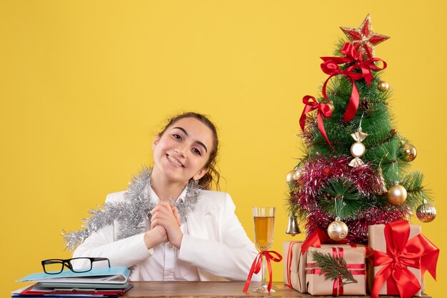 Ärztin der Vorderansicht, die hinter Tabelle auf gelbem Hintergrund mit Weihnachtsbaum und Geschenkboxen sitzt