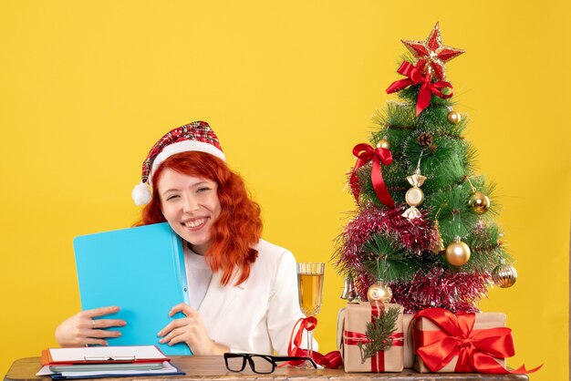 Ärztin der Vorderansicht, die hinter Tabelle sitzt und Dokumente auf gelbem Hintergrund mit Weihnachtsbaum und Geschenkboxen hält