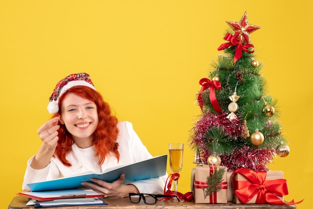 Ärztin der Vorderansicht, die hinter Tabelle sitzt und Dokumente auf gelbem Hintergrund mit Weihnachtsbaum und Geschenkboxen liest