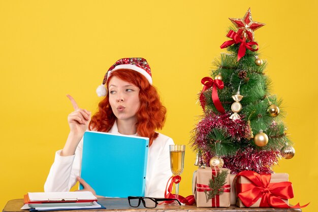 Ärztin der Vorderansicht, die hinter Tisch mit Dokumenten in ihren Händen auf gelbem Hintergrund mit Weihnachtsbaum und Geschenkboxen sitzt