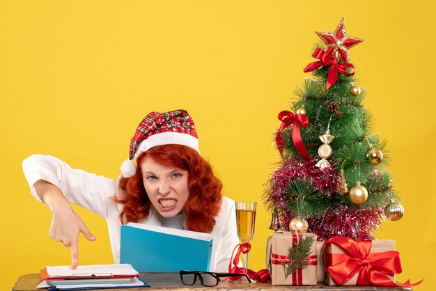 Ärztin der Vorderansicht, die hinter Tisch mit Dokumenten in ihren Händen auf gelbem Hintergrund mit Weihnachtsbaum und Geschenkboxen sitzt