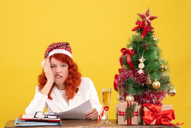 Ärztin der Vorderansicht, die hinter Tisch mit Geschenken und Dokumenten auf gelbem Hintergrund mit Weihnachtsbaum und Geschenkboxen sitzt