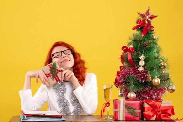 Ärztin der Vorderansicht, die hinter Tisch mit Weihnachtsgeschenken auf gelbem Hintergrund mit Weihnachtsbaum und Geschenkboxen sitzt