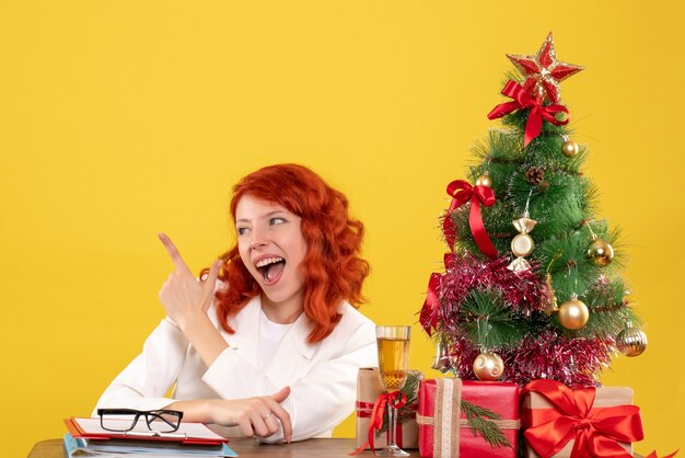 Ärztin der Vorderansicht, die hinter Tisch mit Weihnachtsgeschenken auf gelbem Hintergrund sitzt