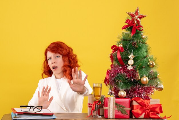 Ärztin der Vorderansicht, die hinter Tisch mit Weihnachtsgeschenken auf gelbem Hintergrund sitzt