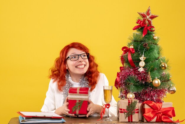 Ärztin der Vorderansicht, die hinter Tisch mit Weihnachtsgeschenken auf gelbem Hintergrund sitzt