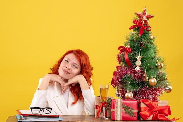 Ärztin der Vorderansicht, die hinter Tisch mit Weihnachtsgeschenken auf gelbem Hintergrund sitzt