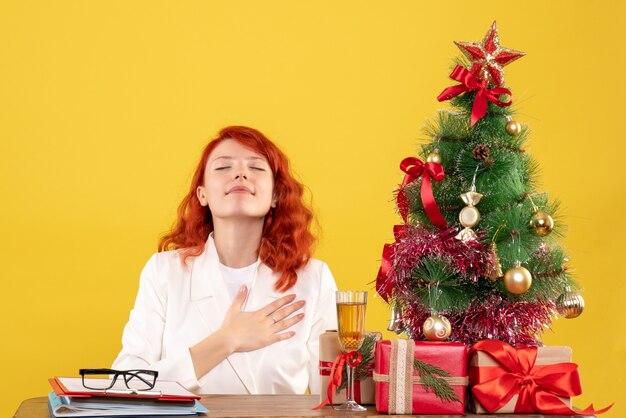 Ärztin der Vorderansicht, die hinter Tisch mit Weihnachtsgeschenken auf gelbem Hintergrund sitzt