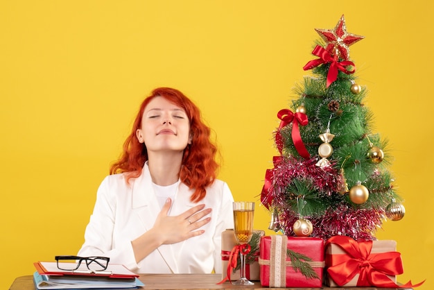 Ärztin der Vorderansicht, die hinter Tisch mit Weihnachtsgeschenken auf gelbem Hintergrund sitzt