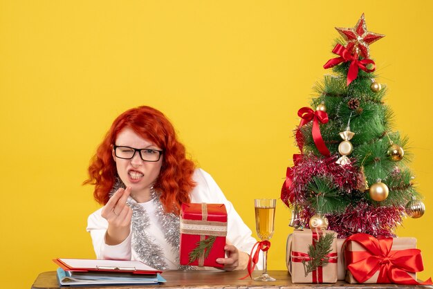 Ärztin der Vorderansicht, die hinter Tisch mit Weihnachtsgeschenken auf gelbem Hintergrund sitzt
