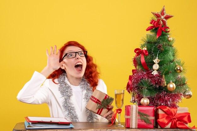 Ärztin der Vorderansicht, die hinter Tisch mit Weihnachtsgeschenken auf gelbem Hintergrund sitzt