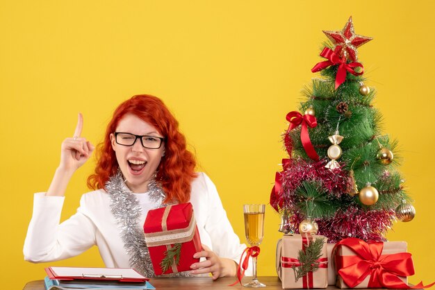 Ärztin der Vorderansicht, die hinter Tisch mit Weihnachtsgeschenken auf gelbem Hintergrund sitzt