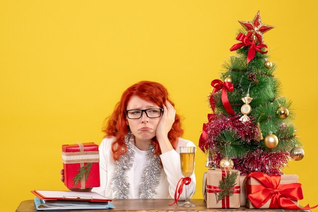 Ärztin der Vorderansicht, die hinter Tisch mit Weihnachtsgeschenken auf gelbem Hintergrund sitzt