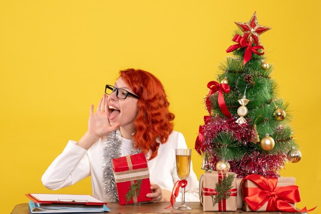 Ärztin der Vorderansicht, die hinter Tisch mit Weihnachtsgeschenken auf gelbem Hintergrund sitzt