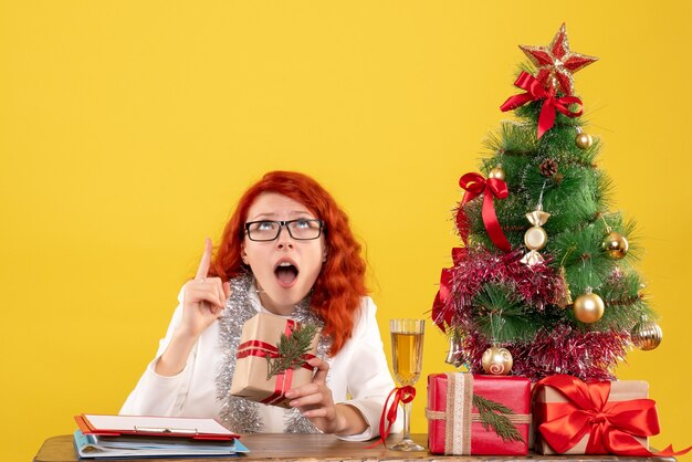 Ärztin der Vorderansicht, die hinter Tisch mit Weihnachtsgeschenken auf gelbem Hintergrund sitzt