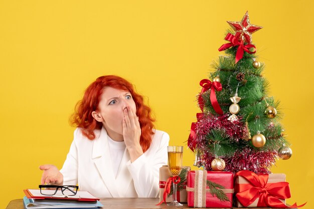 Ärztin der Vorderansicht, die hinter Tisch mit Weihnachtsgeschenken auf gelbem Hintergrund sitzt