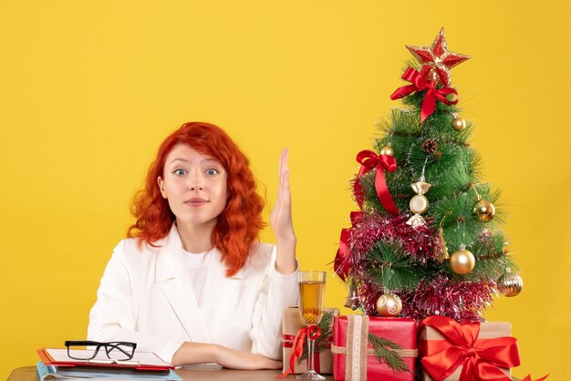 Ärztin der Vorderansicht, die hinter Tisch mit Weihnachtsgeschenken auf gelbem Hintergrund sitzt