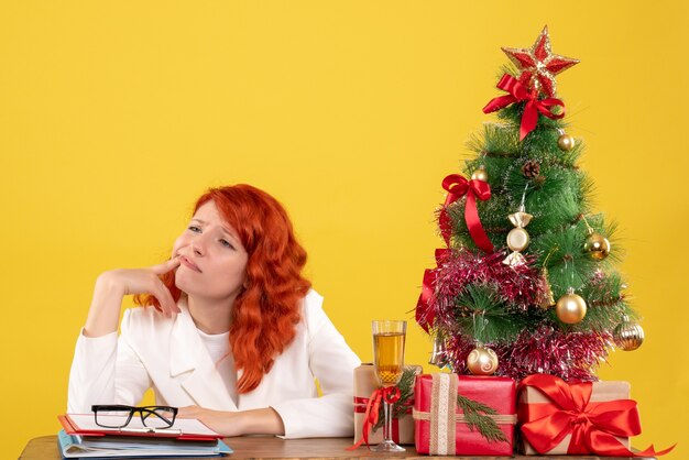 Ärztin der Vorderansicht, die hinter Tisch mit Weihnachtsgeschenken auf gelbem Hintergrund sitzt