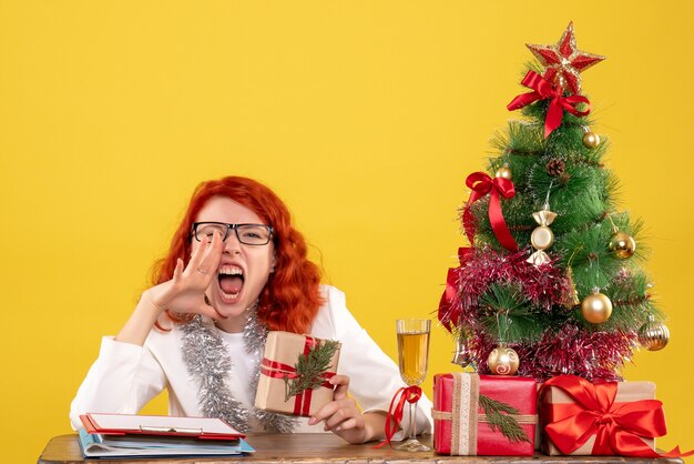 Ärztin der Vorderansicht, die hinter Tisch mit Weihnachtsgeschenken sitzt, die auf gelbem Hintergrund schreien