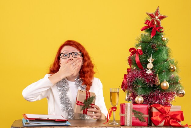 Ärztin der Vorderansicht, die hinter Tisch mit Weihnachtsgeschenken sitzt, schockiert auf gelbem Hintergrund