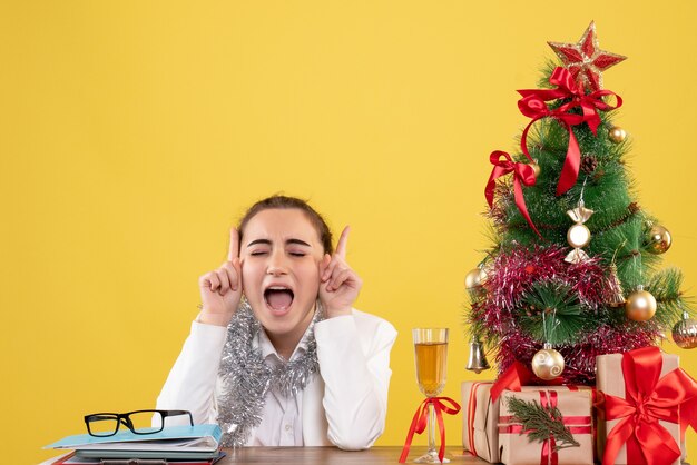 Ärztin der Vorderansicht, die hinter Tisch mit Weihnachtsgeschenken und Baum auf gelbem Hintergrund sitzt