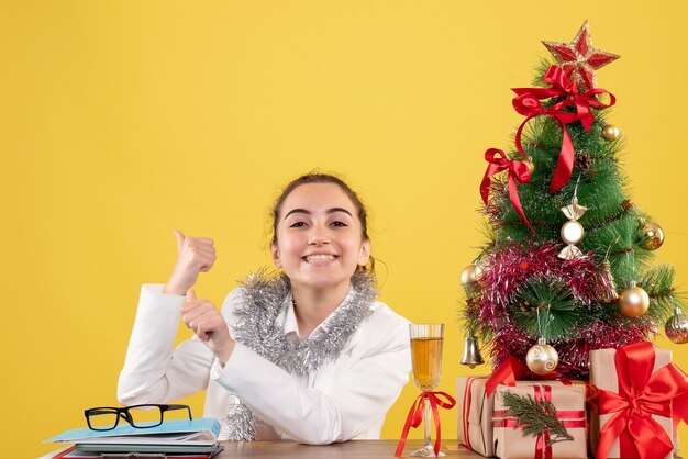 Ärztin der Vorderansicht, die hinter Tisch mit Weihnachtsgeschenken und Baum auf gelbem Hintergrund sitzt