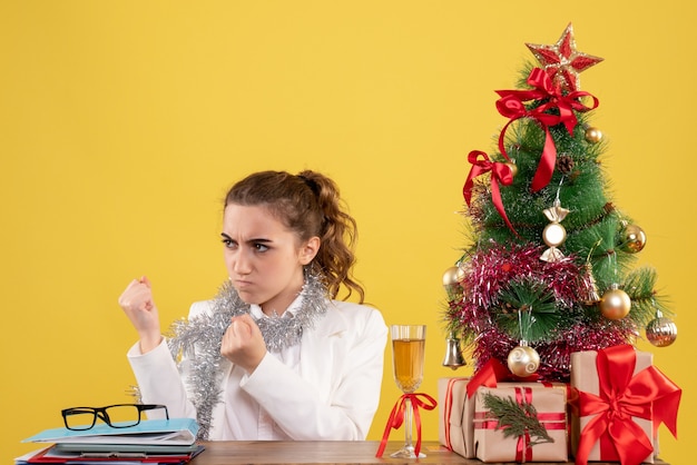 Ärztin der Vorderansicht, die hinter Tisch mit wütendem Gesicht auf gelbem Hintergrund mit Weihnachtsbaum und Geschenkboxen sitzt