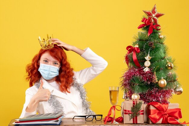 Ärztin der Vorderansicht, die in der Maske mit Weihnachten sitzt, präsentiert das Tragen der Krone auf gelbem Hintergrund