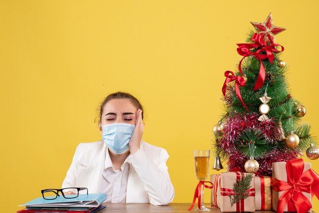 Ärztin der Vorderansicht, die in der Schutzmaske auf gelbem Hintergrund mit Weihnachtsbaum und Geschenkboxen sitzt