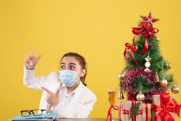 Ärztin der Vorderansicht, die in der Schutzmaske auf gelbem Hintergrund mit Weihnachtsbaum und Geschenkboxen sitzt