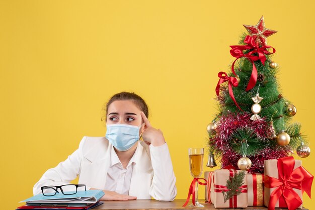 Ärztin der Vorderansicht, die in der Schutzmaske auf gelbem Hintergrund mit Weihnachtsbaum und Geschenkboxen sitzt