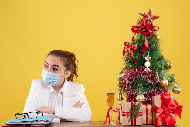 Ärztin der Vorderansicht, die in der Schutzmaske auf gelbem Hintergrund mit Weihnachtsbaum und Geschenkboxen sitzt