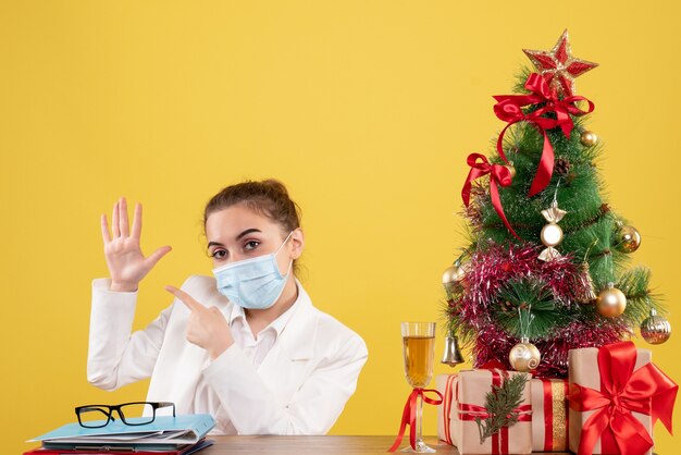 Ärztin der Vorderansicht, die in der Schutzmaske auf gelbem Hintergrund mit Weihnachtsbaum und Geschenkboxen sitzt