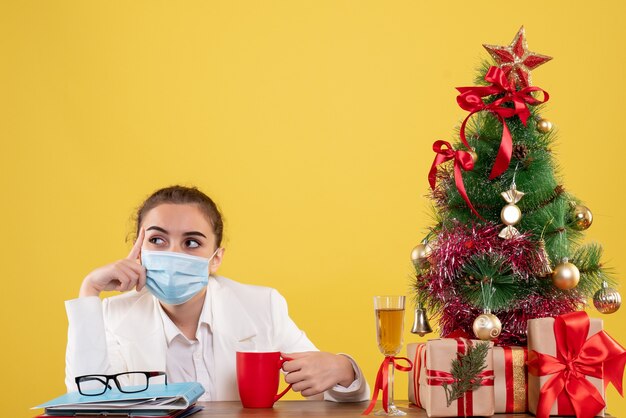 Ärztin der Vorderansicht, die in der Schutzmaske auf gelbem Hintergrund mit Weihnachtsbaum und Geschenkboxen sitzt