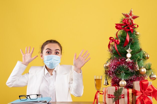 Ärztin der Vorderansicht, die in der Schutzmaske auf gelbem Hintergrund mit Weihnachtsbaum und Geschenkboxen sitzt