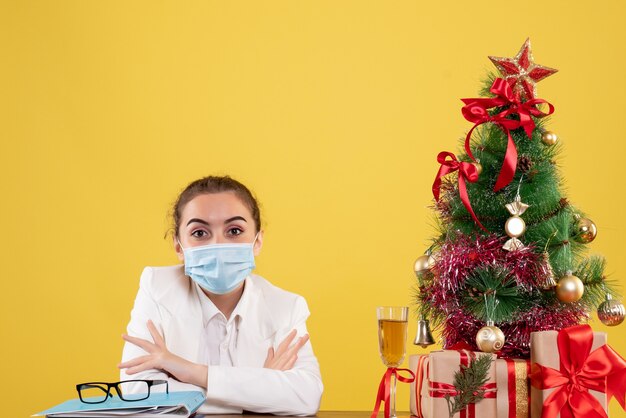 Ärztin der Vorderansicht, die in der Schutzmaske auf gelbem Hintergrund mit Weihnachtsbaum und Geschenkboxen sitzt