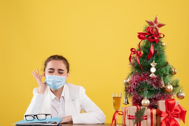 Ärztin der Vorderansicht, die in der Schutzmaske auf gelbem Hintergrund mit Weihnachtsbaum und Geschenkboxen sitzt