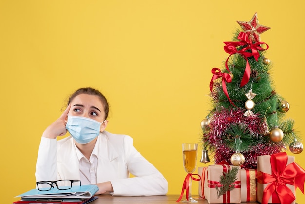 Ärztin der Vorderansicht, die in der sterilen Maske auf gelbem Hintergrund mit Weihnachtsbaum und Geschenkboxen sitzt