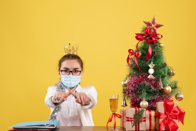 Ärztin der Vorderansicht, die in der sterilen Maske auf gelbem Hintergrund mit Weihnachtsbaum und Geschenkboxen sitzt