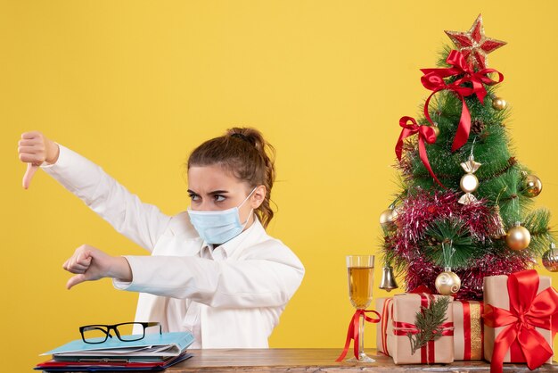 Ärztin der Vorderansicht, die in der sterilen Maske auf gelbem Hintergrund mit Weihnachtsbaum und Geschenkboxen sitzt