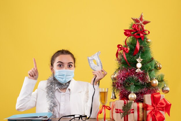 Ärztin der Vorderansicht, die in der sterilen Maske auf gelbem Hintergrund mit Weihnachtsbaum und Geschenkboxen sitzt