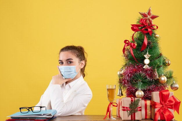 Ärztin der Vorderansicht, die in der sterilen Maske auf gelbem Hintergrund mit Weihnachtsbaum und Geschenkboxen sitzt