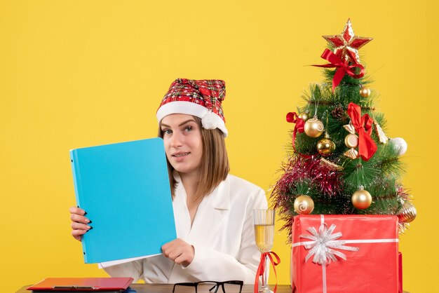 Ärztin der Vorderansicht, die mit Weihnachten sitzt, präsentiert das Halten von Dateien auf gelbem Hintergrund