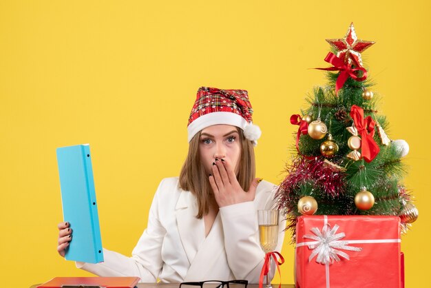 Ärztin der Vorderansicht, die mit Weihnachten sitzt, präsentiert das Halten von Dokumenten auf gelbem Hintergrund