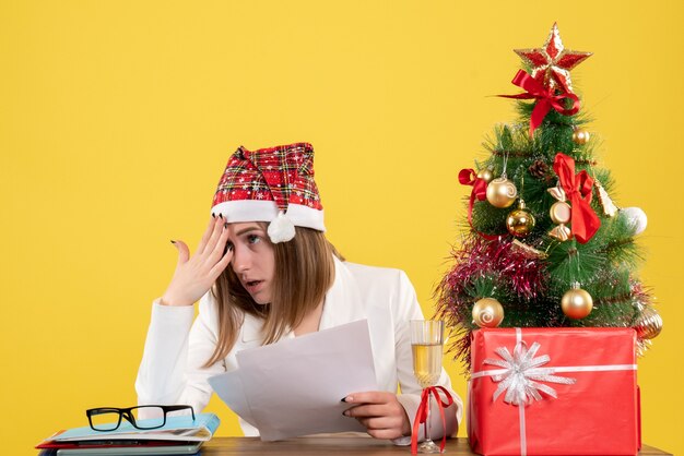 Ärztin der Vorderansicht, die mit Weihnachten sitzt, präsentiert das Halten von Dokumenten auf gelbem Hintergrund